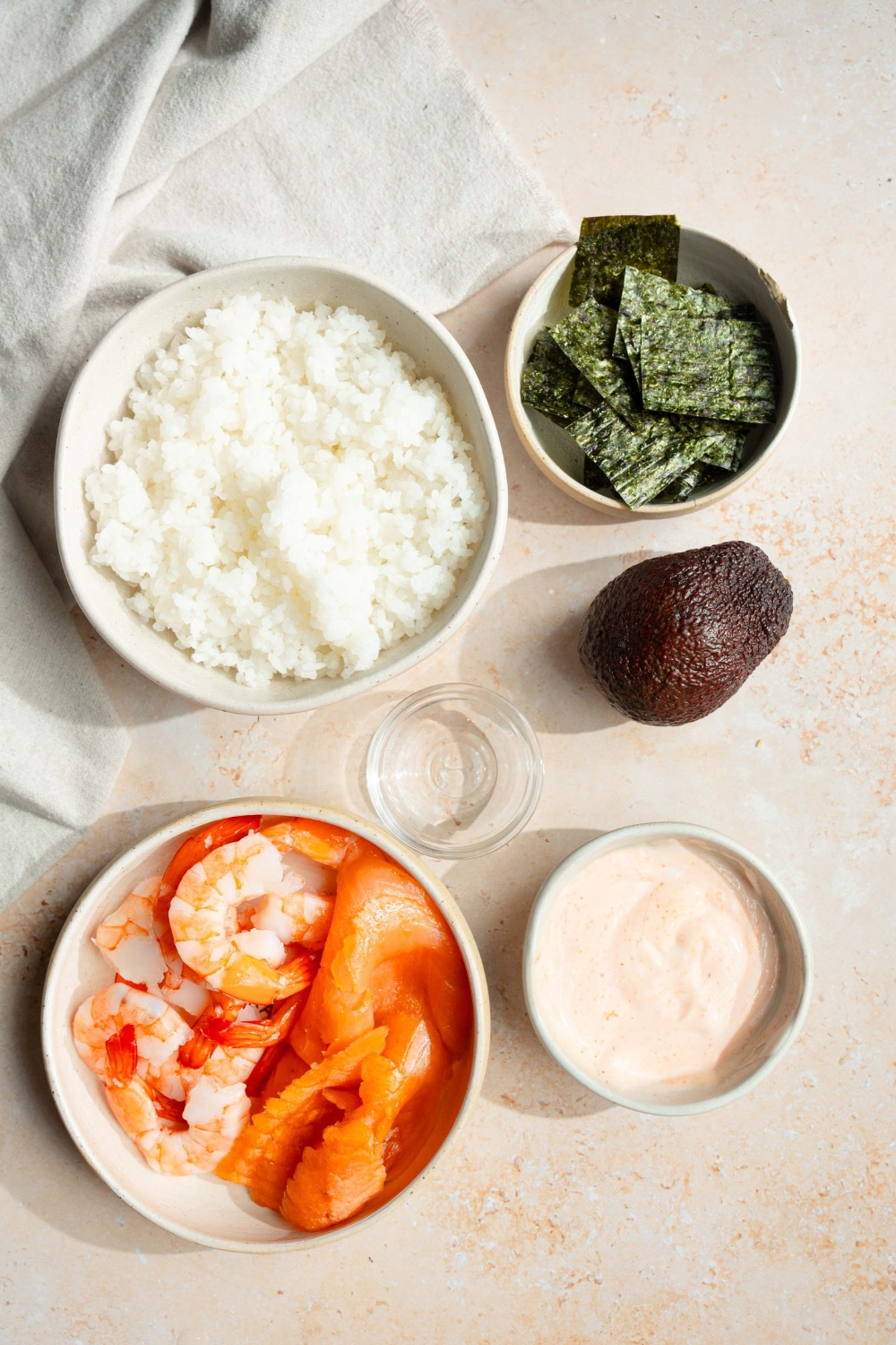 A bowl of rice, a bowl of Nori sheets, a bowl of shrimp and avocado, and a bowl of pie mayo all on a counter