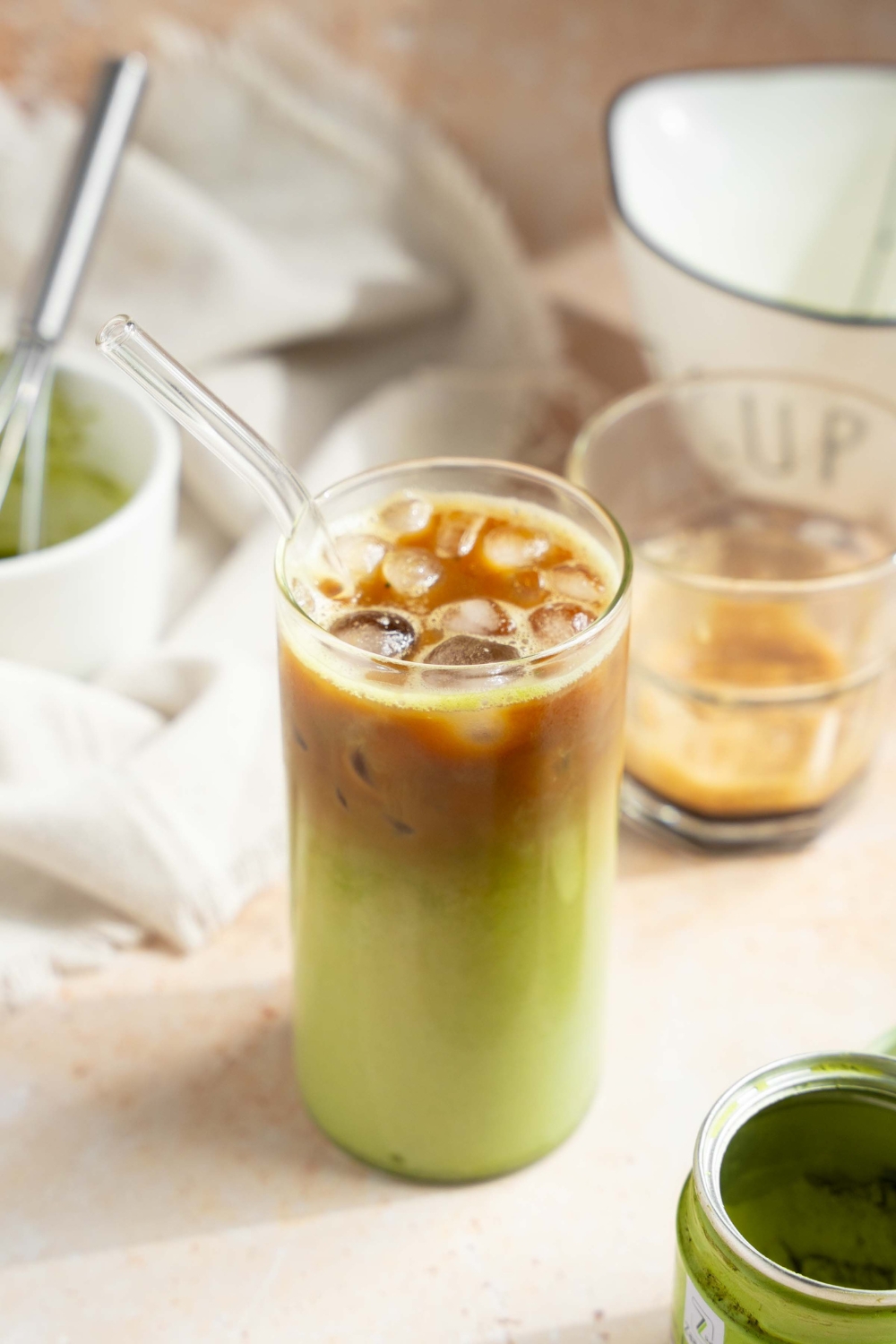 A tall glass of iced matcha latte with a glass straw. The glass is on a tan counter with a tin of matcha powder, small glass of poured espresso, and a small bowl and whisk with matcha.