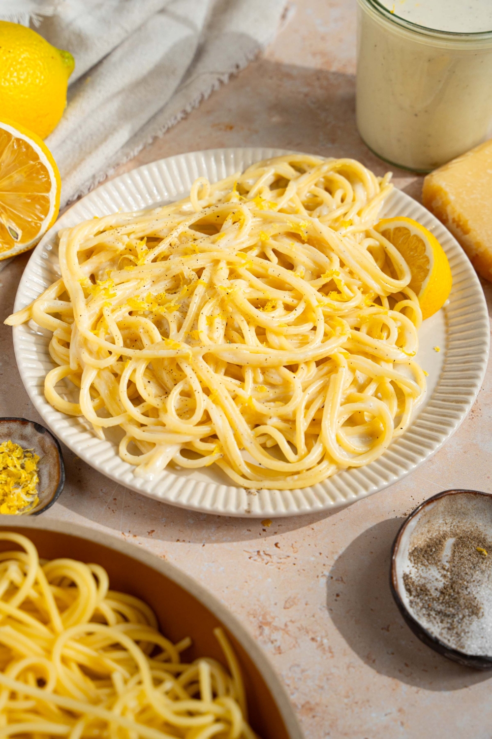 A white plate with spaghetti tossed in lemon cream sauce garnished with black pepper and served with a lemon wedge. The plate is on a tan counter with a bowl of pasta, small bowl of garnishes, and sliced lemons.