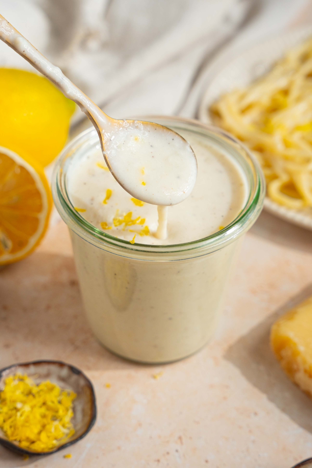 A glass jar with lemon cream sauce garnished with lemon zest. There is a spoon dipping into the jar. The jar is on a tan counter with a plate of pasta, halved lemons, and small bowl of lemon zest.