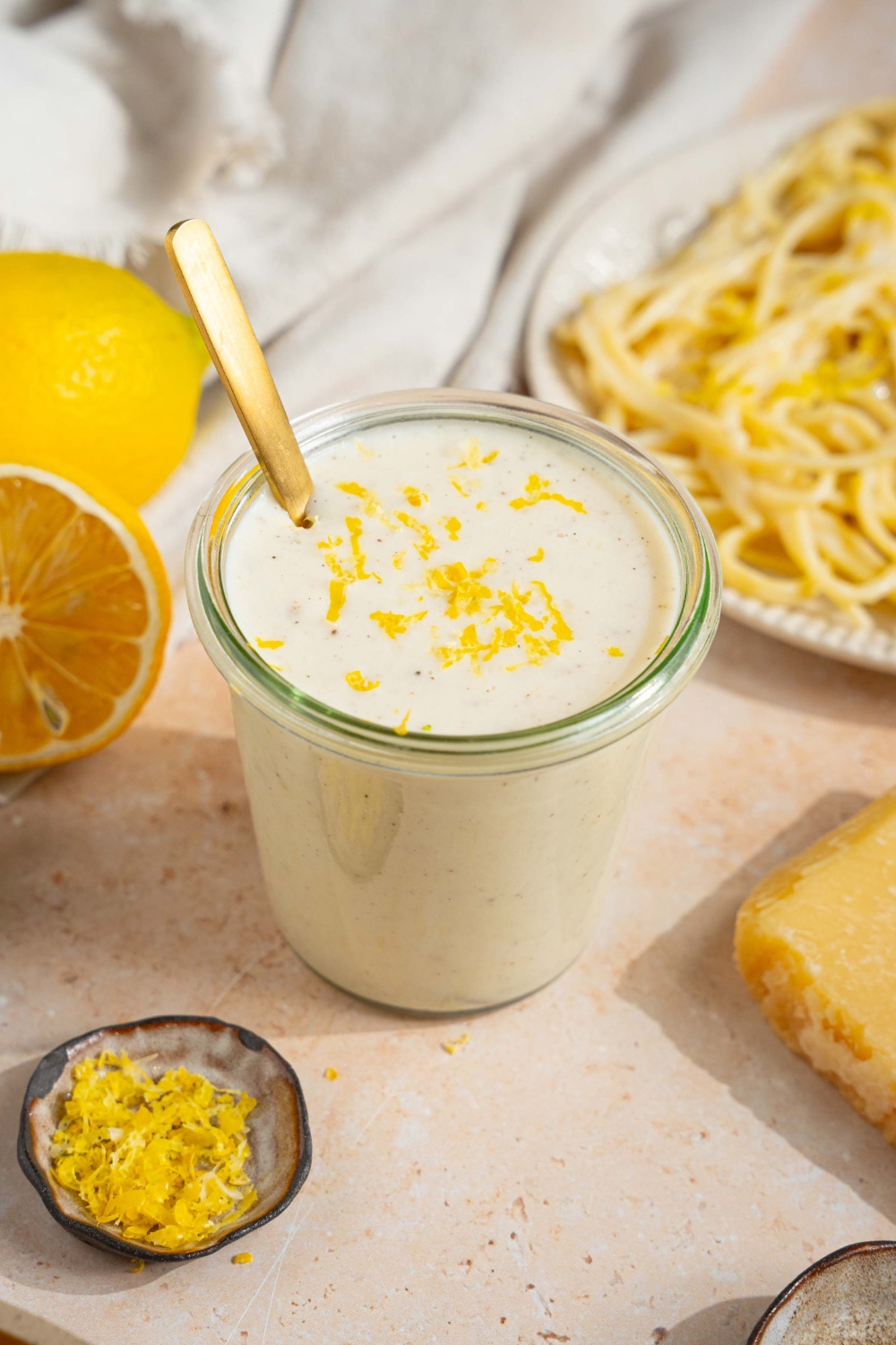 A glass jar with lemon cream sauce garnished with lemon zest. There is a spoon in the jar. The jar is on a tan counter with a plate of pasta, halved lemons, and small bowl of lemon zest.