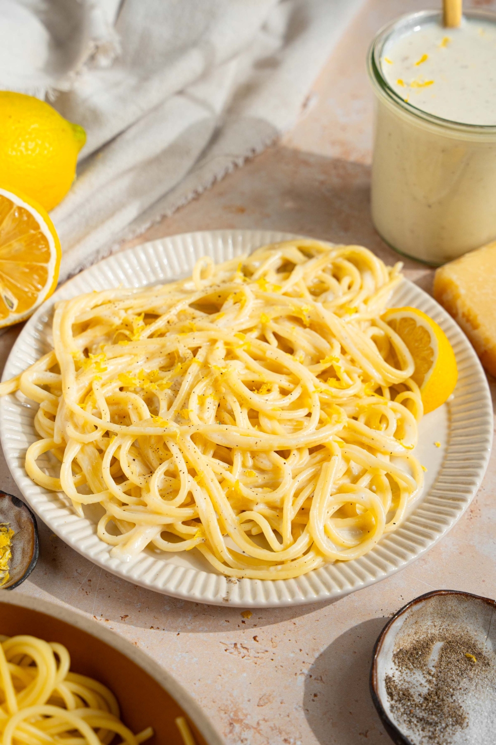 A white plate with spaghetti tossed in lemon cream sauce garnished with black pepper and served with a lemon wedge. The plate is on a tan counter with a jar of cream sauce, small bowl of garnishes, and halved lemons.