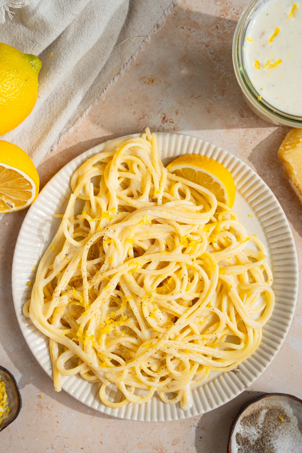 A white plate with spaghetti tossed in lemon cream sauce garnished with black pepper and served with a lemon wedge. The plate is on a tan counter with a jar of cream sauce, small bowl of garnishes, and halved lemons.