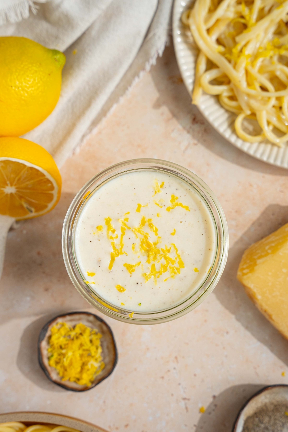 A glass jar with lemon cream sauce garnished with lemon zest. The jar is on a tan counter with a plate of pasta, halved lemons, and small bowl of lemon zest.