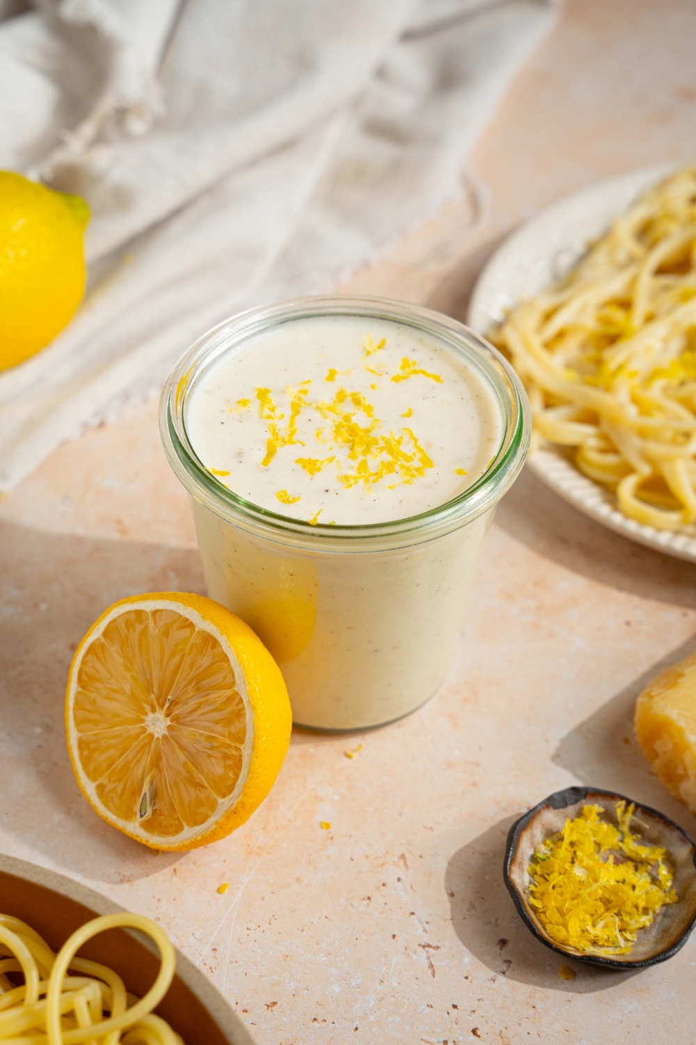 A glass jar with lemon cream sauce garnished with lemon zest. The jar is on a tan counter with a plate of pasta, halved lemons, and small bowl of lemon zest.