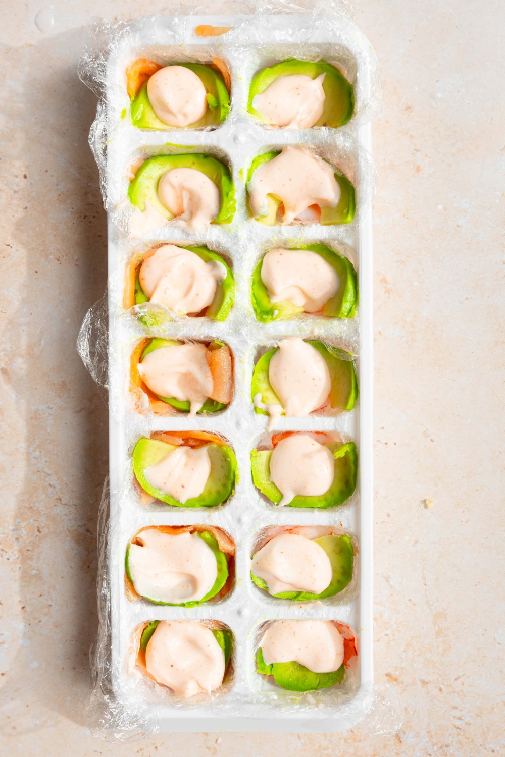 An ice cube tray that has mayo on top of avocado that is on shrimp.