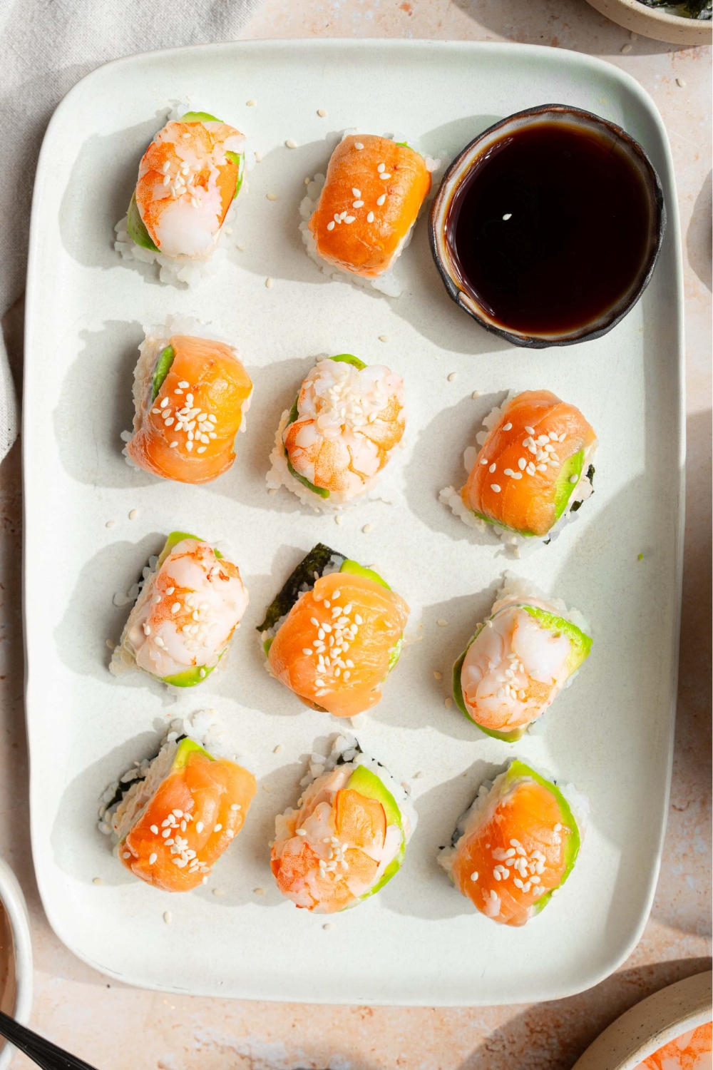 11 pieces of sushi lined up in rose on a white tray. The back row has two pieces of sushi and on the right hand side of them as a cup of soy sauce.