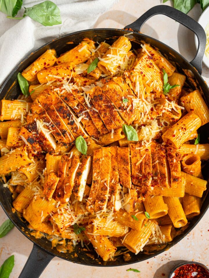 A skillet with garlic parmesan chicken pasta garnished with fresh bail, shredded parmesan and red pepper flakes. The skillet is on a tan counter with a white cloth napkin and small plate of garnishes.