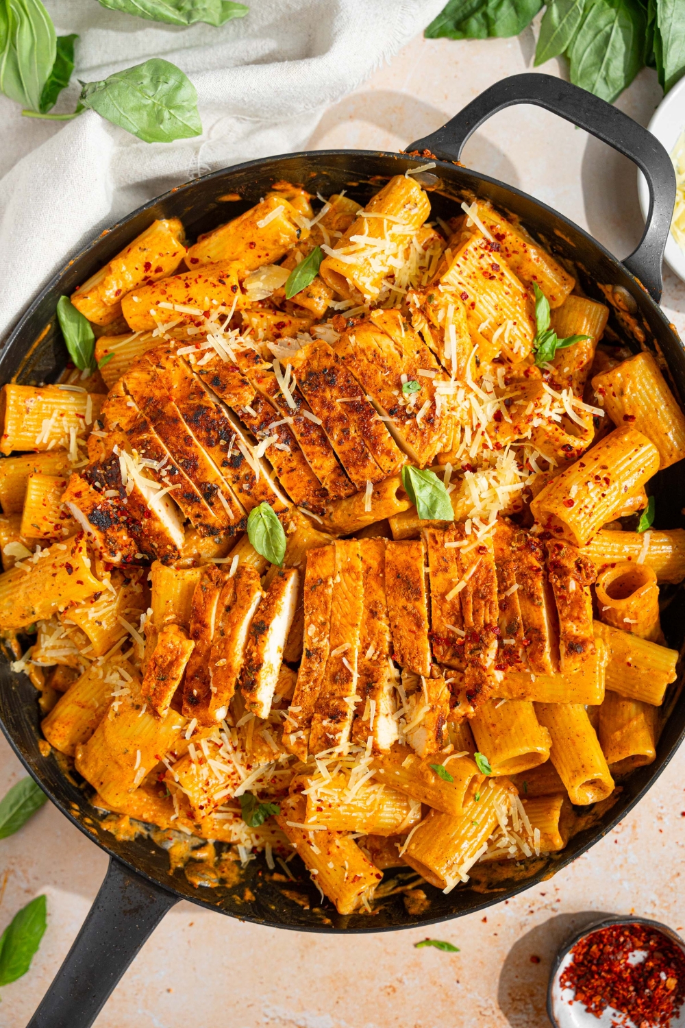 A skillet with garlic parmesan chicken pasta garnished with fresh bail, shredded parmesan and red pepper flakes. The skillet is on a tan counter with a white cloth napkin and small plate of garnishes.