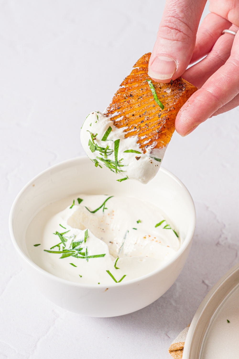 A hand dipping a seasoned baked accordion potato into a bowl of dip. The dip is garnished with fresh parsley. The bowl is on a white counter.