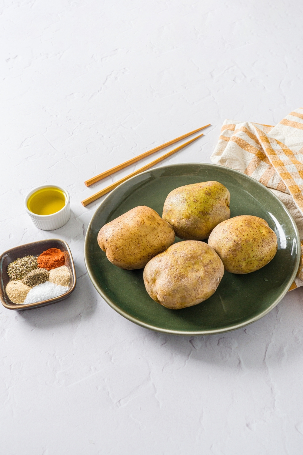 A white counter with ingredients to make accordion potatoes including white potatoes, oil, chopsticks, and seasonings.