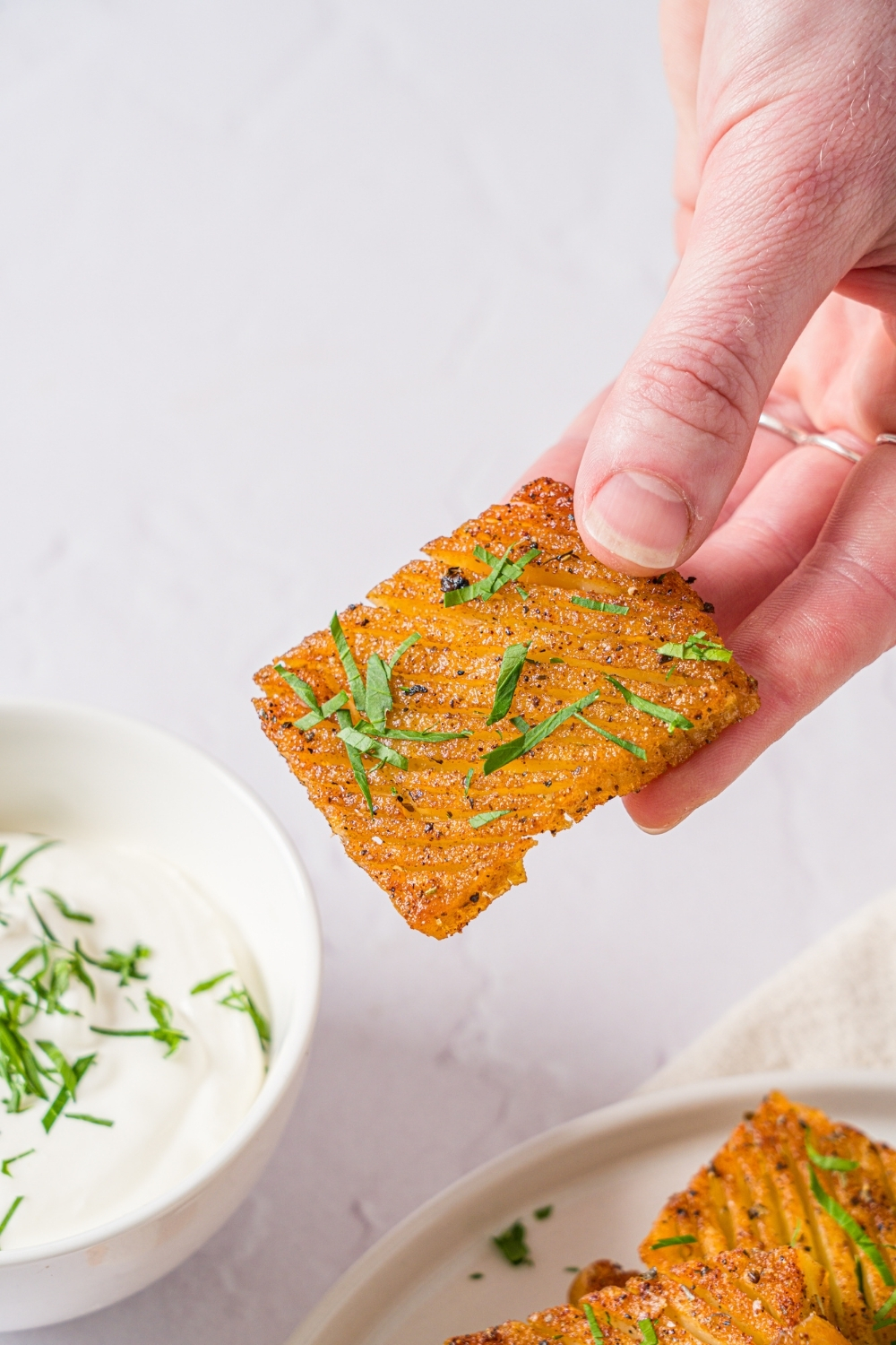 A hand holding a seasoned baked accordion potato garnished with fresh parsley. There is a white counter with a plate of potatoes and bowl of dip in the background.
