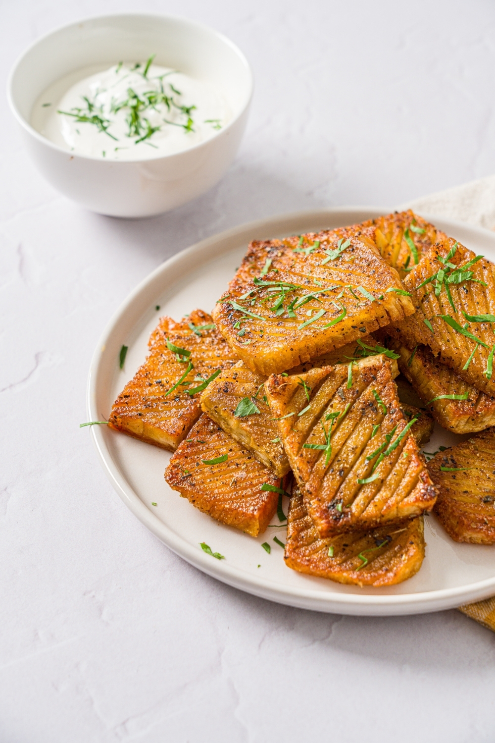 A white plate with seasoned baked accordion potatoes garnished with fresh parsley. The plate is on a white counter with a bowl of dip garnished with fresh parsley.