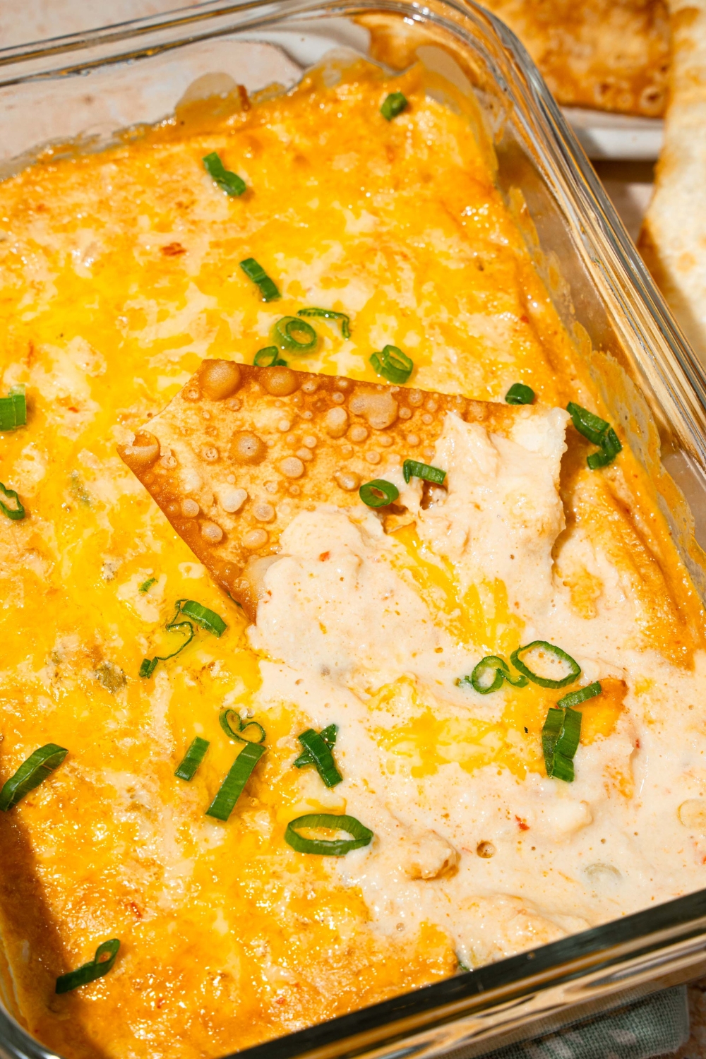 A glass baking dish with baked crab rangoon dip garnished with sliced green onions. A wonton chip is in the dip.