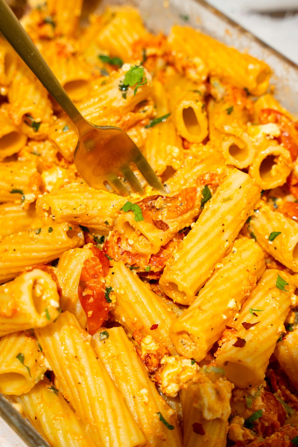 A glass baking dish with baked feta pasta garnished with fresh parsley and crushed red pepper. A fork is in the dish.