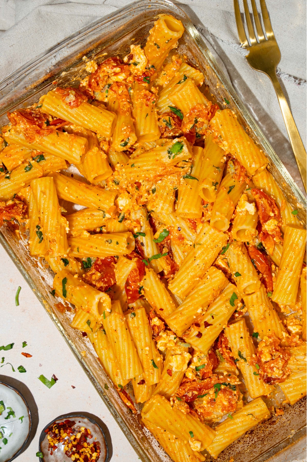 A glass baking dish with baked feta pasta garnished with fresh parsley and crushed red pepper. The dish is on a tan counter with a fork and small bowls of garnishes.