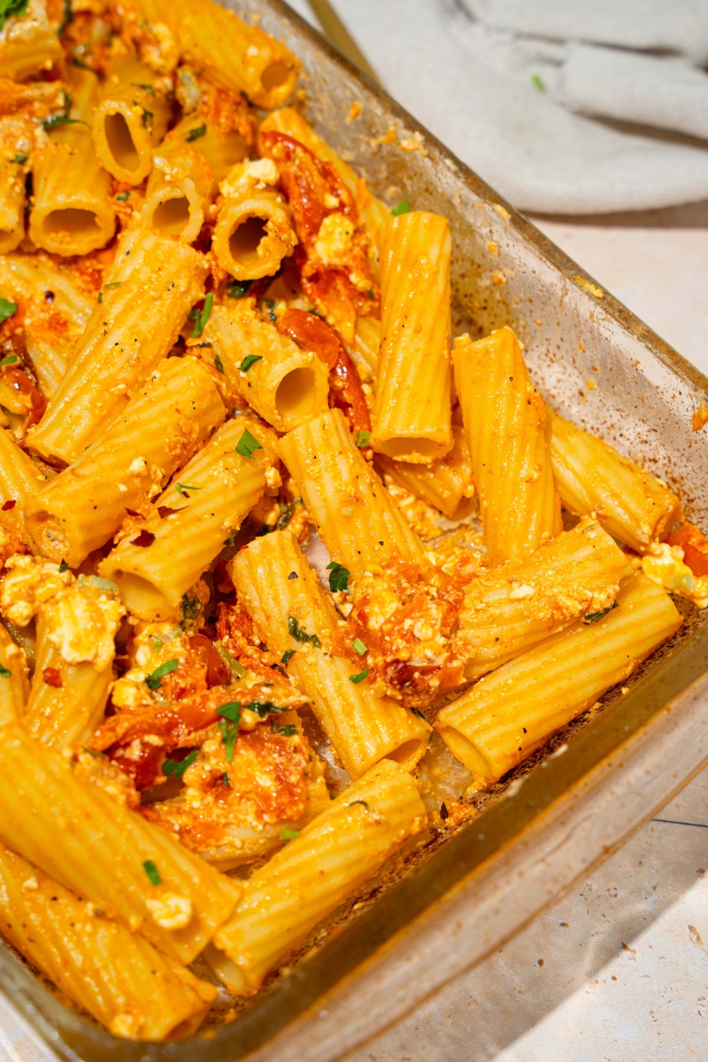A glass baking dish with baked feta pasta garnished with fresh parsley and crushed red pepper. The dish is on a tan counter with a white cloth napkin.