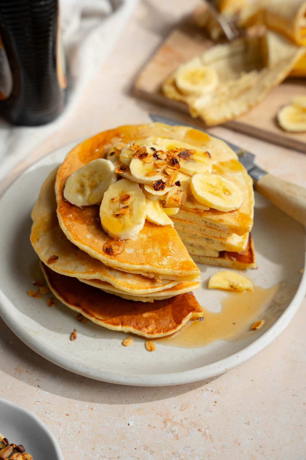 A white plate with a stack of fluffy banana pancakes topped with sliced banana and granola drizzled with syrup. There is a bite taken from the stack. The plate is on a tan counter with a bottle of syrup and wooden cutting board with bananas.