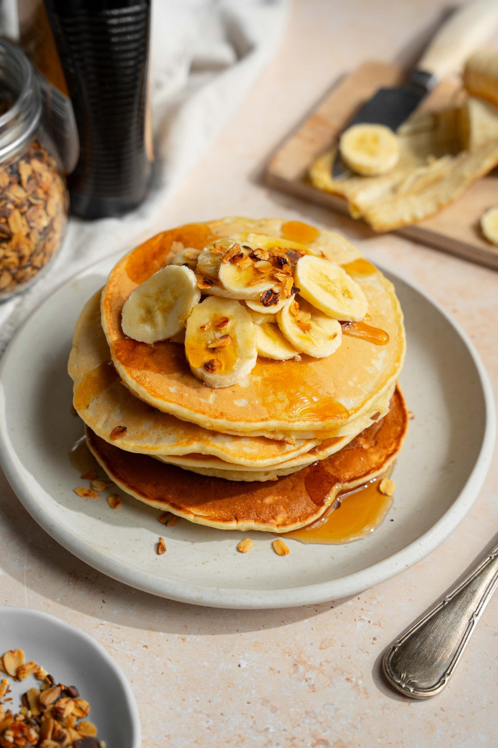A white plate with a stack of fluffy banana pancakes topped with sliced banana and granola drizzled with syrup. The plate is on a tan counter with a bottle of syrup and wooden cutting board with bananas.