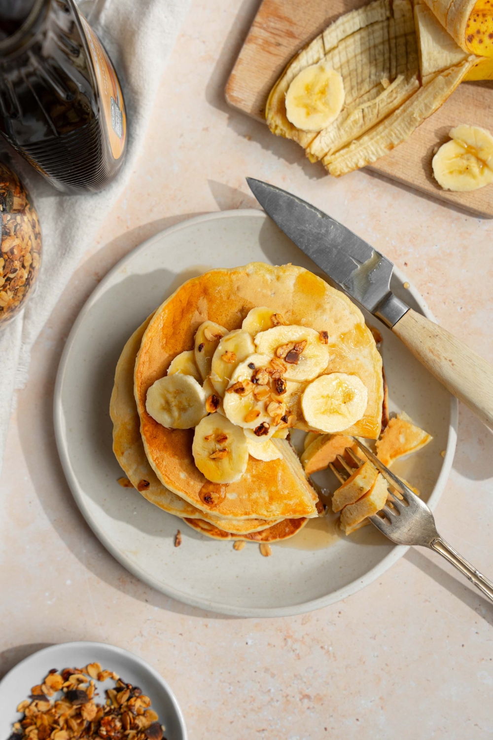 A white plate with a stack of fluffy banana pancakes topped with sliced banana and granola drizzled with syrup. A fork is taking a bite from the stack. The plate is on a tan counter with a knife and wooden cutting board with bananas.