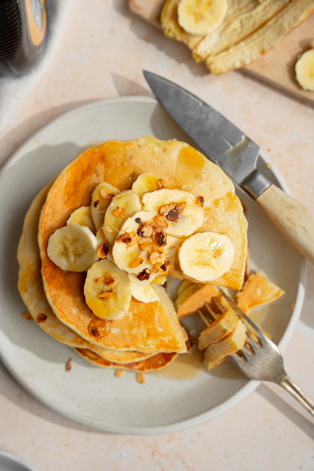 A white plate with a stack of fluffy banana pancakes topped with sliced banana and granola drizzled with syrup. A fork is taking a from the stack. The plate is on a tan counter with a knife and a wooden cutting board with bananas.