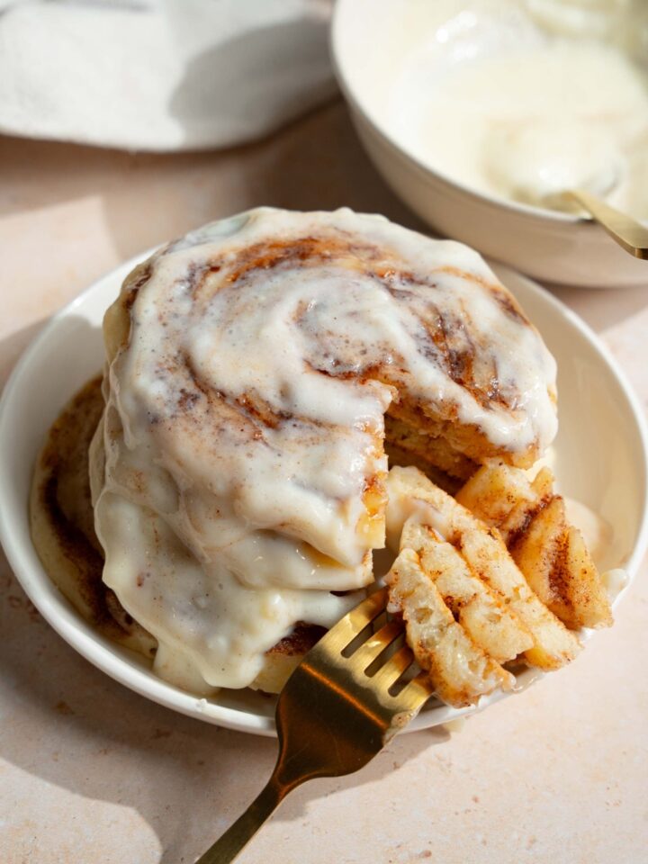 A plate with a stack of cinnamon roll pancakes topped with cream cheese icing. There is a fork with a bite taken from the stack. The plate is on a tan counter with a bowl of cream cheese filling.