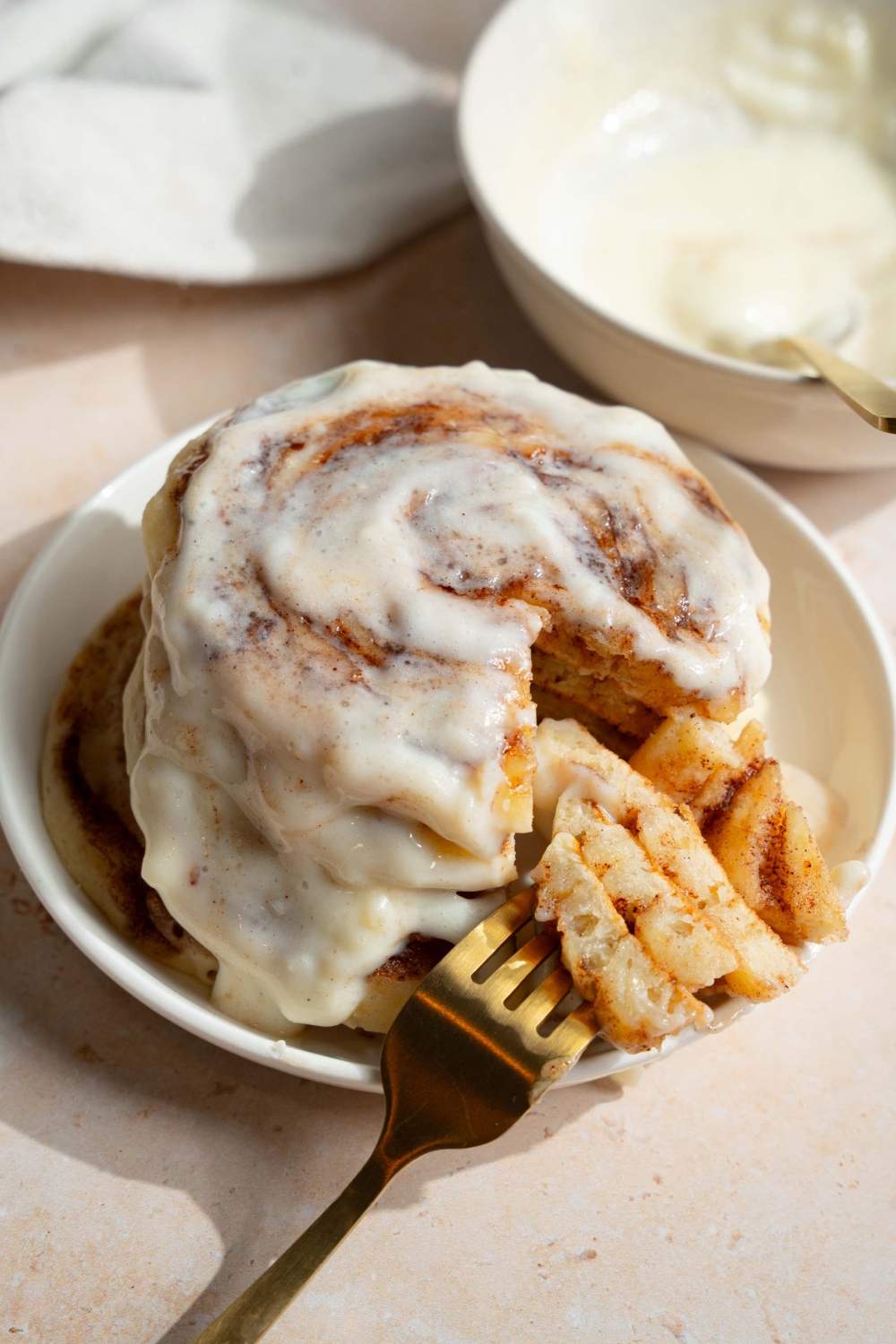 A plate with a stack of cinnamon roll pancakes topped with cream cheese icing. There is a fork with a bite taken from the stack. The plate is on a tan counter with a bowl of cream cheese filling.