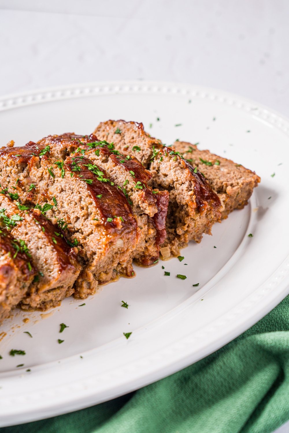 A white platter with sliced meatloaf without eggs. The meatloaf is garnished with fresh parsley. The plate is on a marble counter with a green cloth napkin.