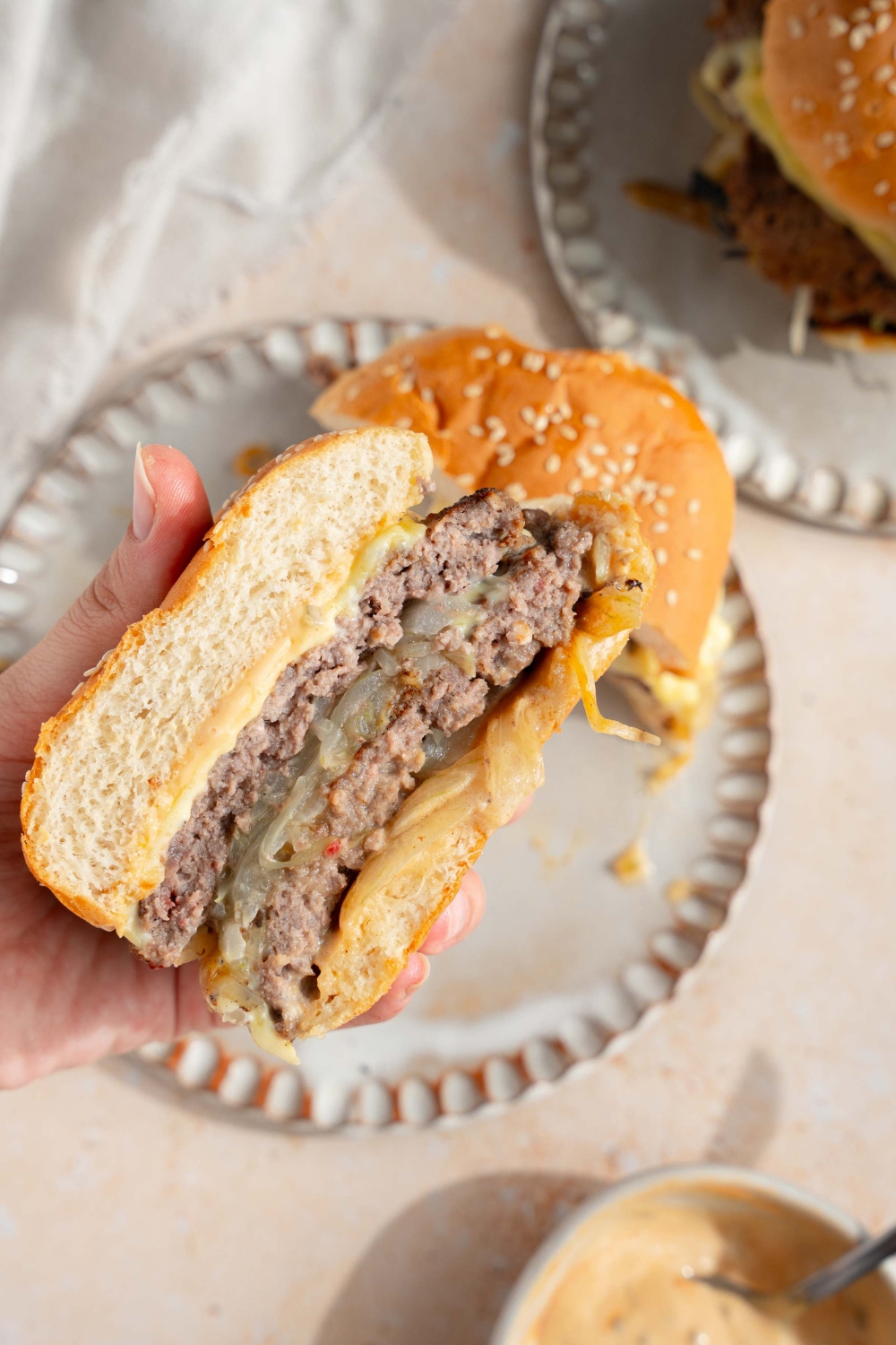 A hand holding a smash burger with onions sliced in half. There is a plate with the other half of the burger. The plate is on a tan counter with an additional smash burger.