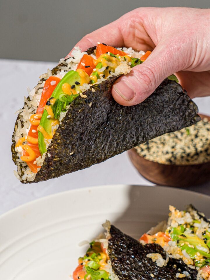 A hand holding a sushi taco made with salmon, sushi rice, avocado, sriracha mayo, and cream cheese wrapped in a nori sheet and garnished with sesame seeds. There is a plate of sushi tacos on a white counter with a bowl of sesame seeds.