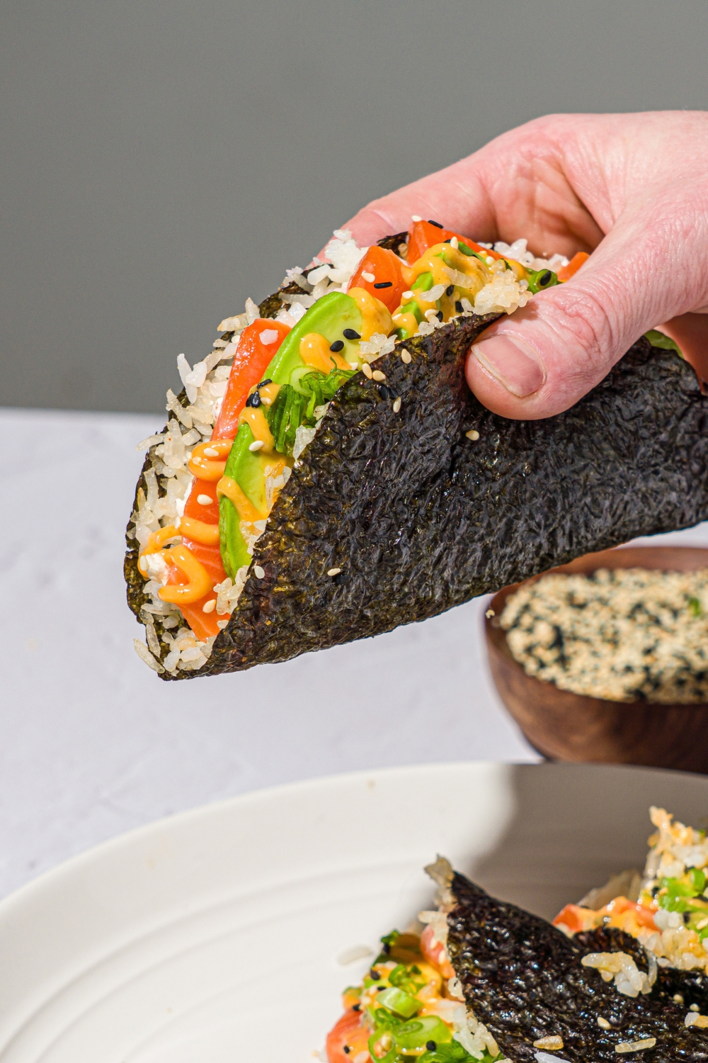 A hand holding a salmon sushi taco made with sushi rice, avocado, sriracha mayo, and cream cheese wrapped in a nori sheet and garnished with sesame seeds. There is a plate of sushi tacos on a white counter with a bowl of sesame seeds.