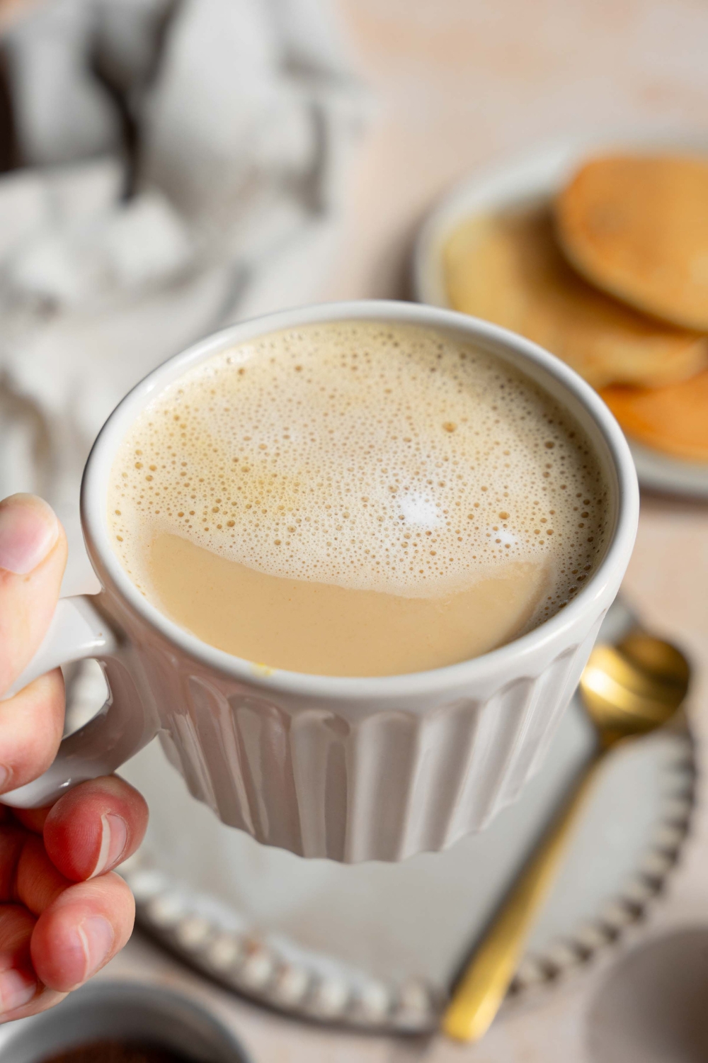 A hand holding a mug of Vietnamese egg coffee. There is a saucer and spoon on a tan counter with a white cloth napkin blurred in the background.