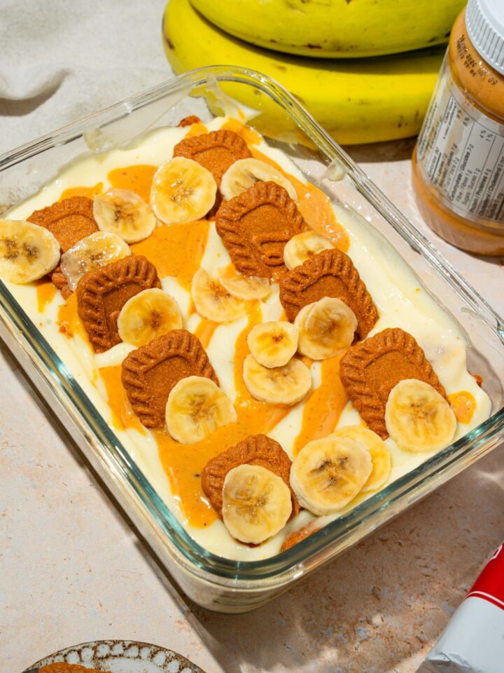 A glass baking dish with Biscoff banana pudding topped with Biscoff cookie pieces, sliced bananas, and caramel drizzle. The dish is on a tan counter with bananas and Biscoff cookies.