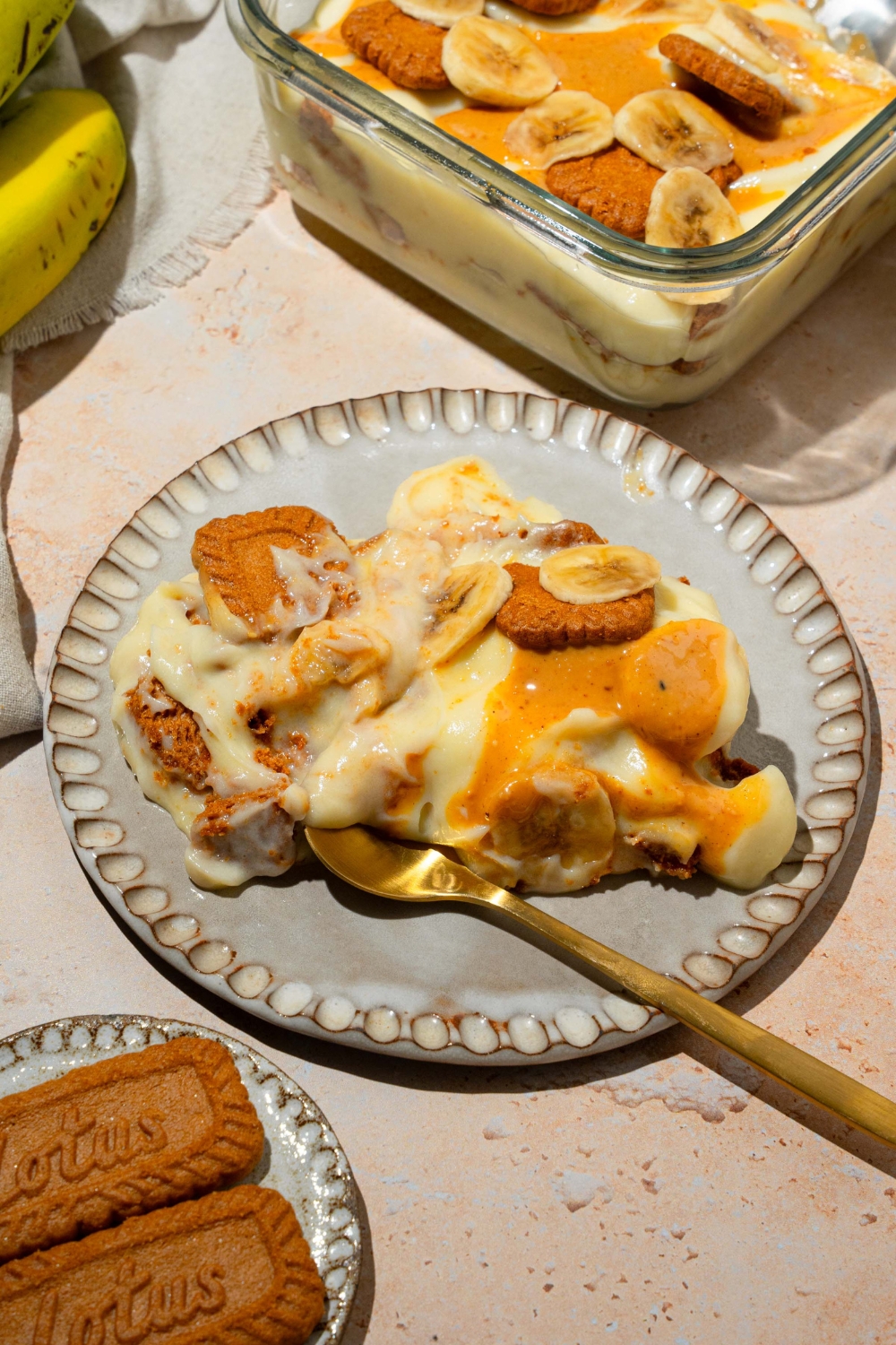 A plate with Biscoff banana pudding drizzled with caramel and topped with Biscoff cookies and sliced banana. There is a spoon in the pudding. The plate is on a tan counter with a baking dish of pudding.
