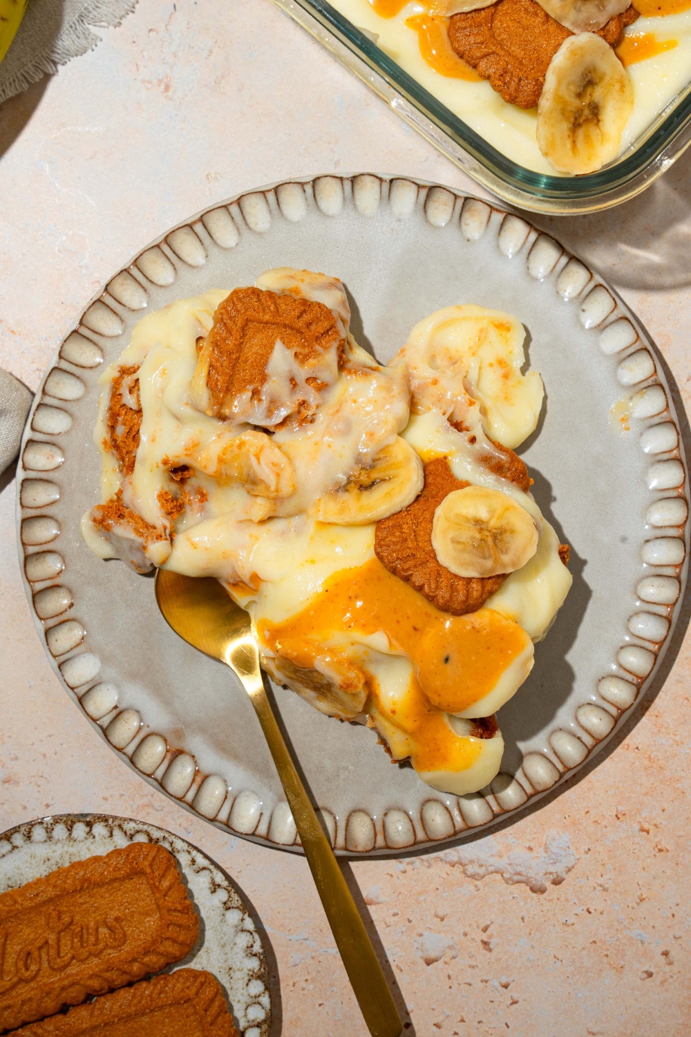 A plate with Biscoff banana pudding drizzled with caramel and topped with Biscoff cookies and sliced banana. There is a spoon in the pudding. The plate is on a tan counter with a small plate of cookies.