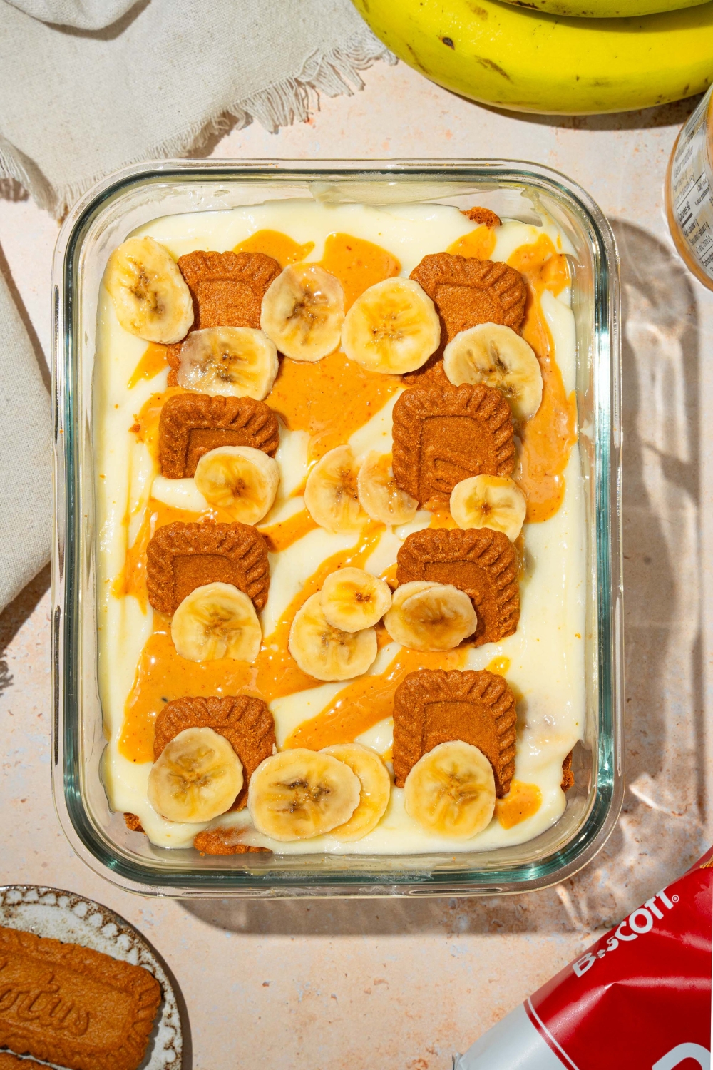 A glass baking dish with Biscoff banana pudding topped with Biscoff cookie pieces, sliced bananas, and caramel drizzle. The dish is on a tan counter with bananas and Biscoff cookies.