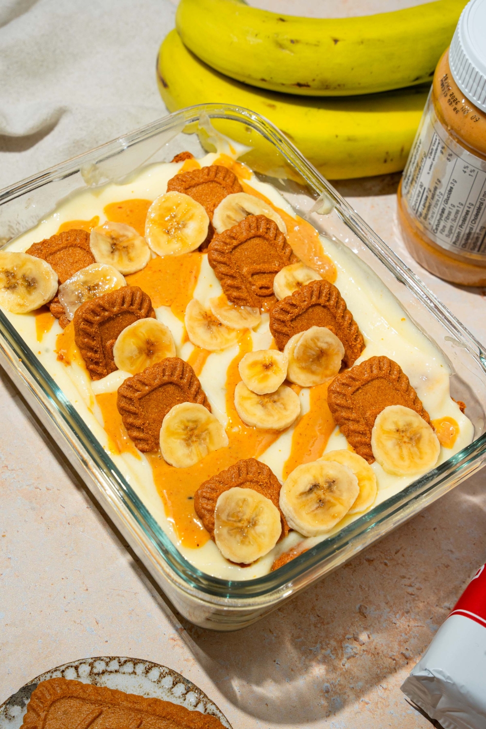 A glass baking dish with Biscoff banana pudding topped with Biscoff cookie pieces, sliced bananas, and caramel drizzle. The dish is on a tan counter with bananas and Biscoff cookies.