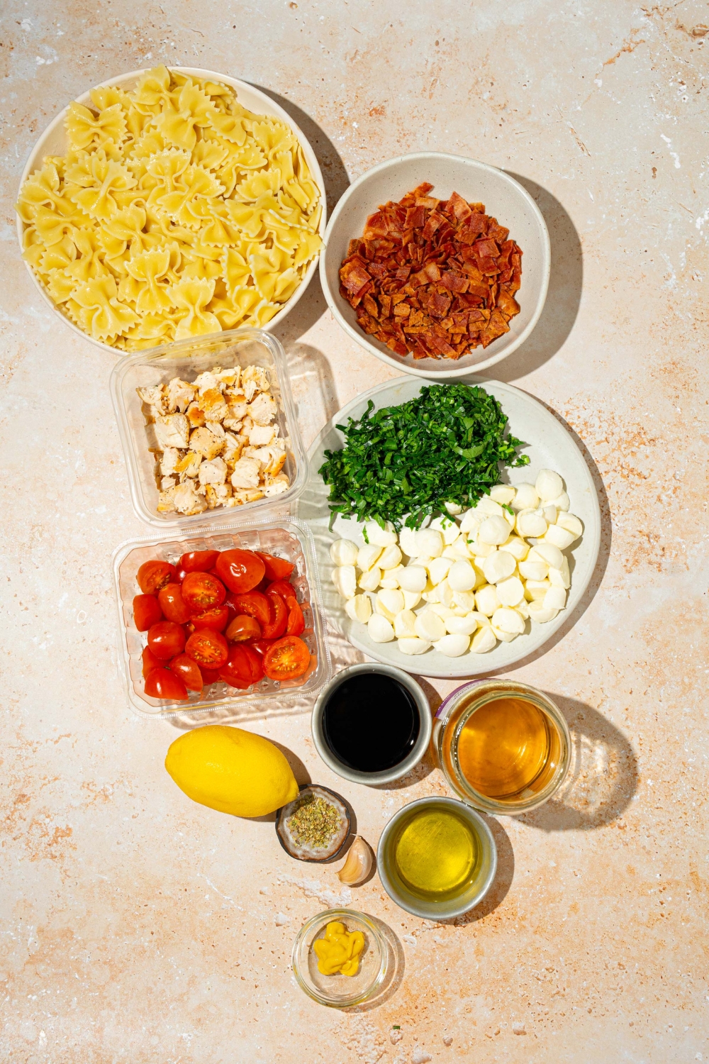 An overhead shot of several bowls in various sizes containing ingredients to make bruschetta chicken pasta including bowtie pasta, grilled chicken, bacon, mozzarella pearls, tomatoes, fresh basil, lemon, balsamic vinegar, honey, olive oil, mustard, and garlic.