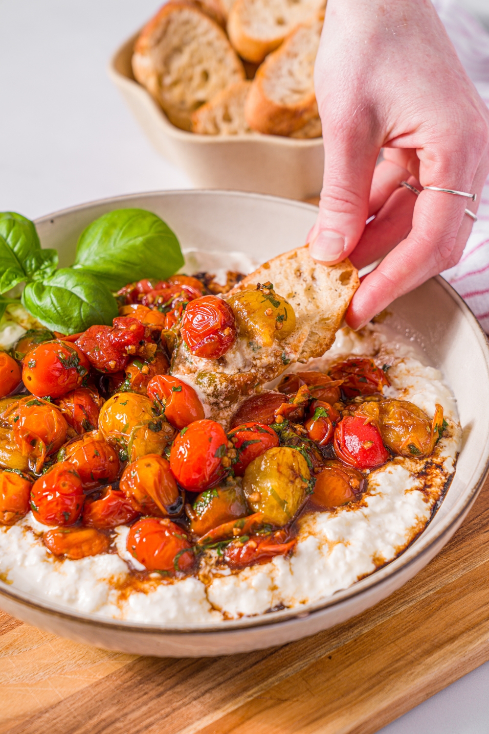 A white bowl with roasted tomato burrata dip garnished with fresh basil. A hand is dipping a toasted crostini into the dip. The bowl is on a wooden board on a marble counter with a small bowl of toasted crostini.
