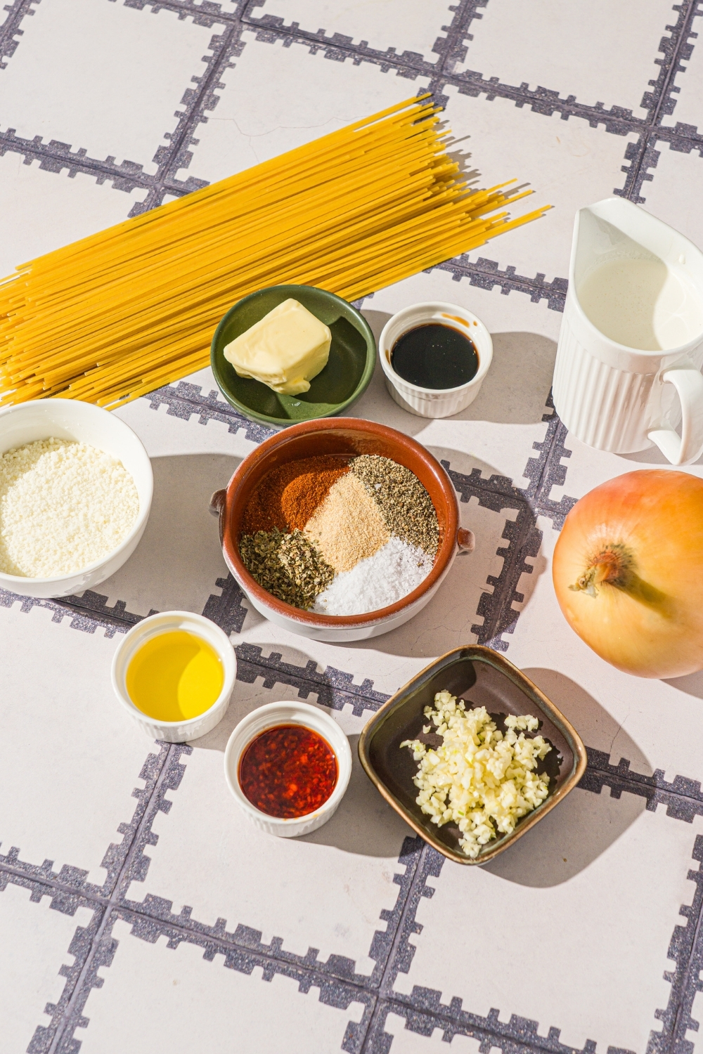 A tiled counter with ingredients to make caramelized onion pasta including spaghetti, onion, garlic, butter, chili crisp oil, soy sauce, olive oil, parmesan cheese, and seasonings.