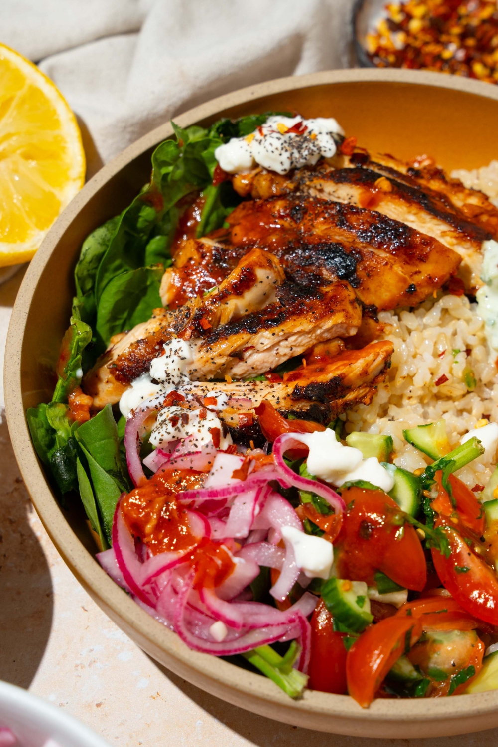 A bowl with Cava mediterranean bowl including rice, sliced marinated chicken, mixed greens, tzatziki, cucumber and tomato salad, and feta cheese in a harissa honey vinaigrette. The bowl is on a tan counter with a lemon and white cloth napkin.