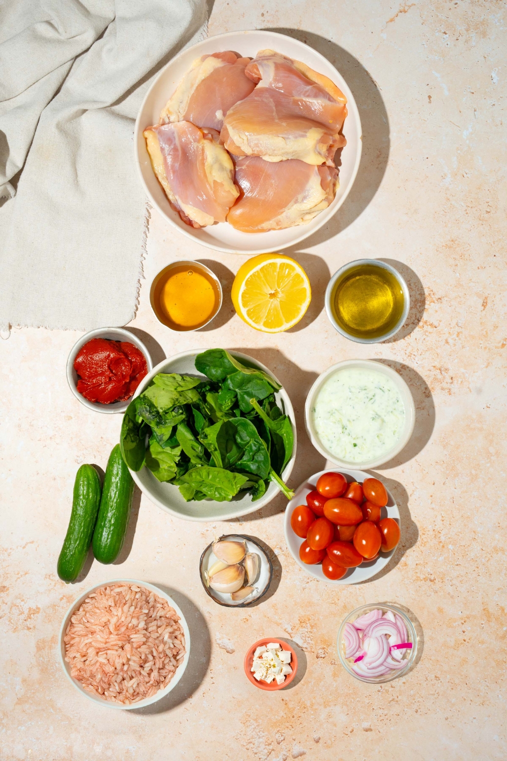 An overhead shot of several bowls in various sizes containing ingredients to make Cava mediterranean bowl incuding chicken, cucumber, tomatoes, harissa paste, honey, lemon, garlic, rice, tzatziki, and more.