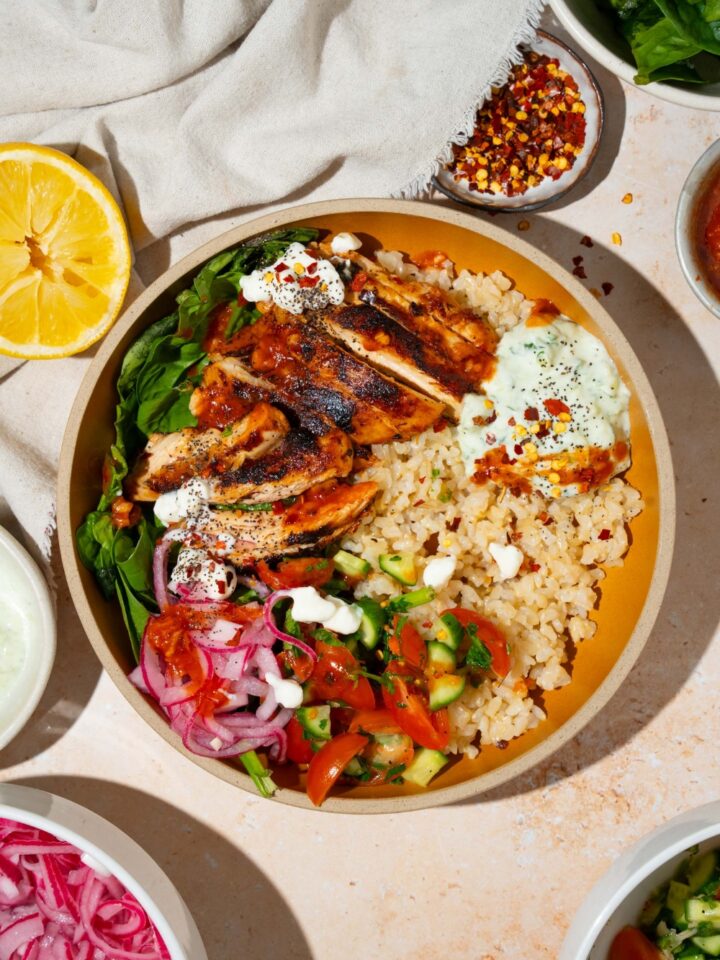 A bowl with Cava mediterranean bowl including rice, sliced marinated chicken, mixed greens, tzatziki, cucumber and tomato salad, and feta cheese in a harissa honey vinaigrette. The bowl is on a tan counter with a small bowls of ingredients and a white cloth napkin.