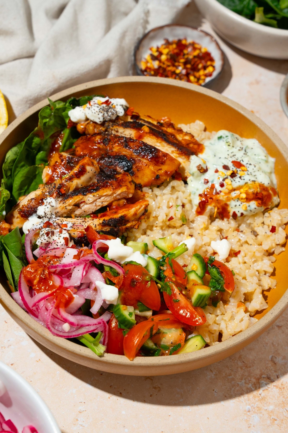 A bowl with Cava mediterranean bowl including rice, sliced marinated chicken, mixed greens, tzatziki, cucumber and tomato salad, and feta cheese in a harissa honey vinaigrette. The bowl is on a tan counter with a small bowl of red pepper flakes and a white cloth napkin.