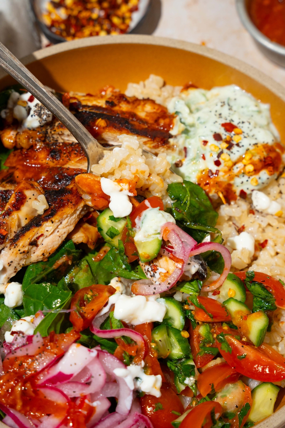 A bowl with Cava mediterranean bowl including rice, sliced marinated chicken, mixed greens, tzatziki, cucumber and tomato salad, and feta cheese in a harissa honey vinaigrette. There is a fork in the bowl.