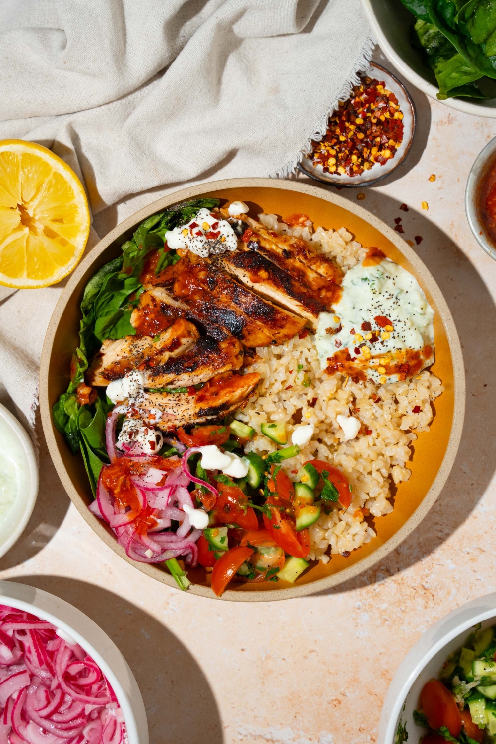 A bowl with Cava mediterranean bowl including rice, sliced marinated chicken, mixed greens, tzatziki, cucumber and tomato salad, and feta cheese in a harissa honey vinaigrette. The bowl is on a tan counter with a small bowls of ingredients and a white cloth napkin.