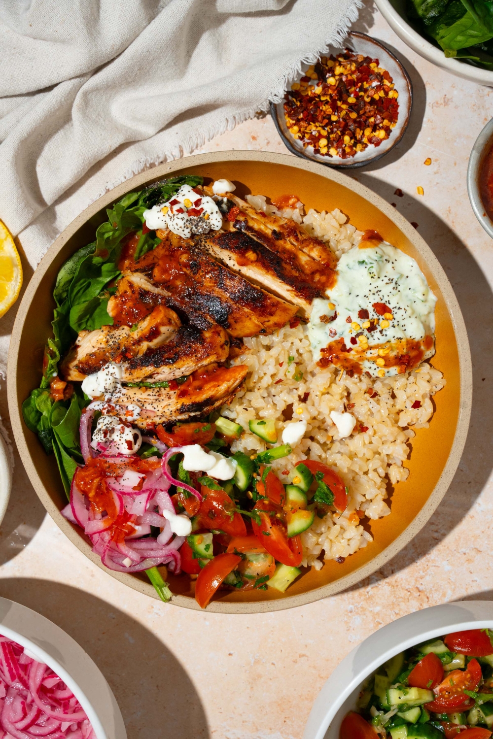 A bowl with Cava mediterranean bowl including rice, sliced marinated chicken, mixed greens, tzatziki, cucumber and tomato salad, and feta cheese in a harissa honey vinaigrette. The bowl is on a tan counter with a small bowls of ingredients and a white cloth napkin.