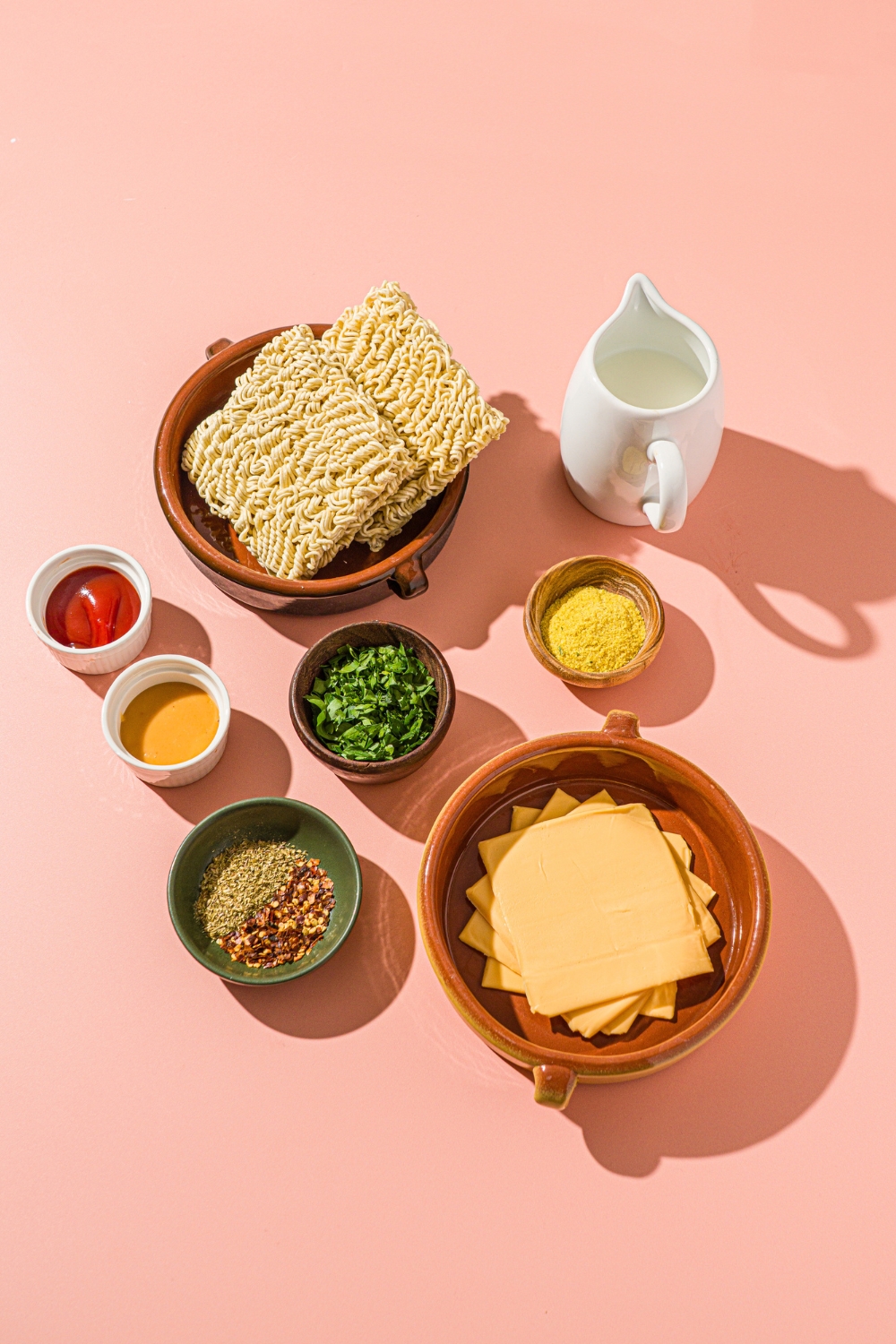 A pink counter with ingredients to make cheesy maggi including maggi noodles, milk, American cheese, Schezwan sauce, seasoned mayo, and seasonings.