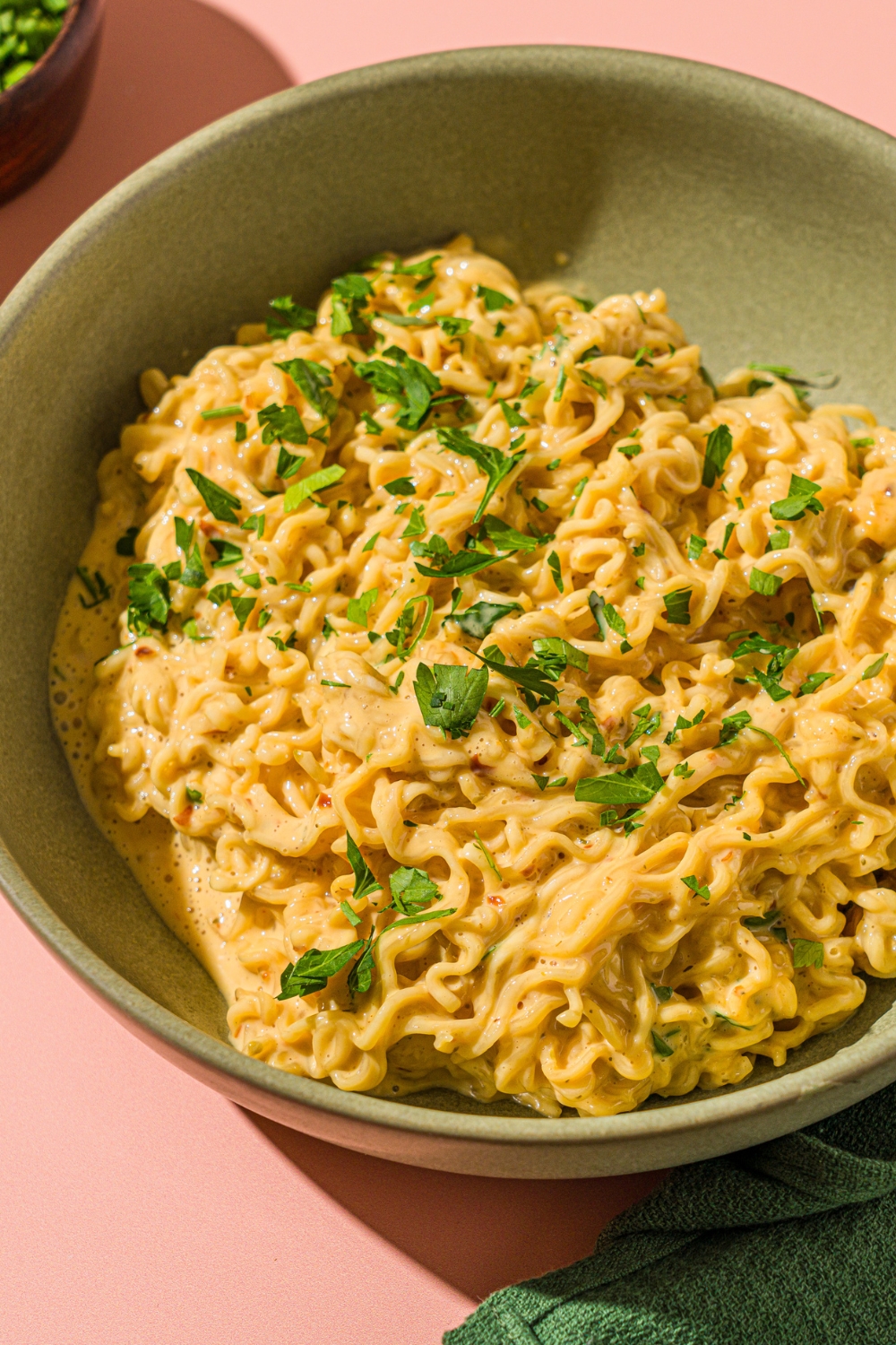 A green bowl with cheesy Maggi noodles garnished with fresh cilantro. The bowl is on a pink counter with a green cloth napkin.