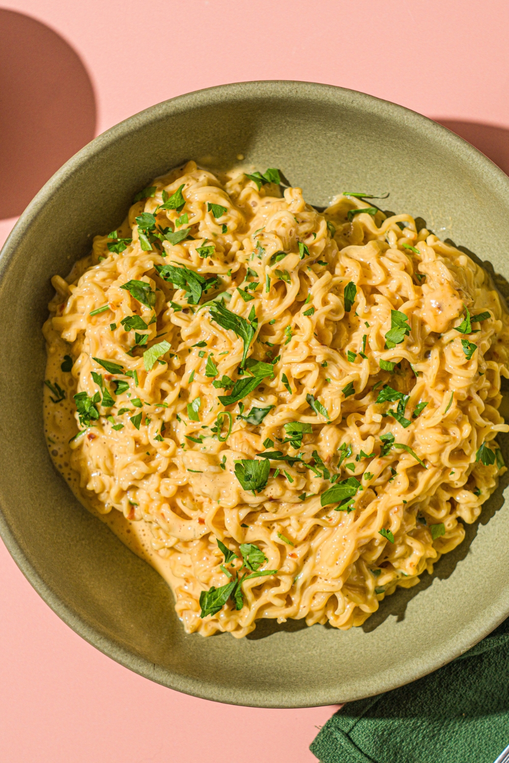 A green bowl with cheesy Maggi noodles garnished with fresh cilantro. The bowl is on a pink counter with a green cloth napkin.